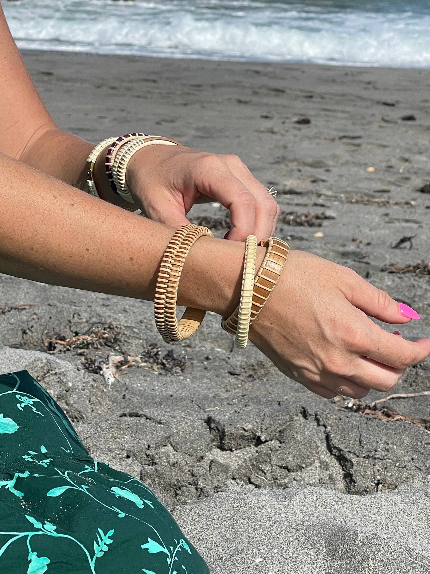 woman wearing a green print dress showing her hands wearing multiple wooden woven bracelets in front of a dark snad beach with small waves