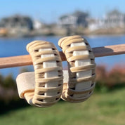 A pair of medium-sized hoop earrings made with woven design, displayed on a wooden stick, with water and buildings in the background.
