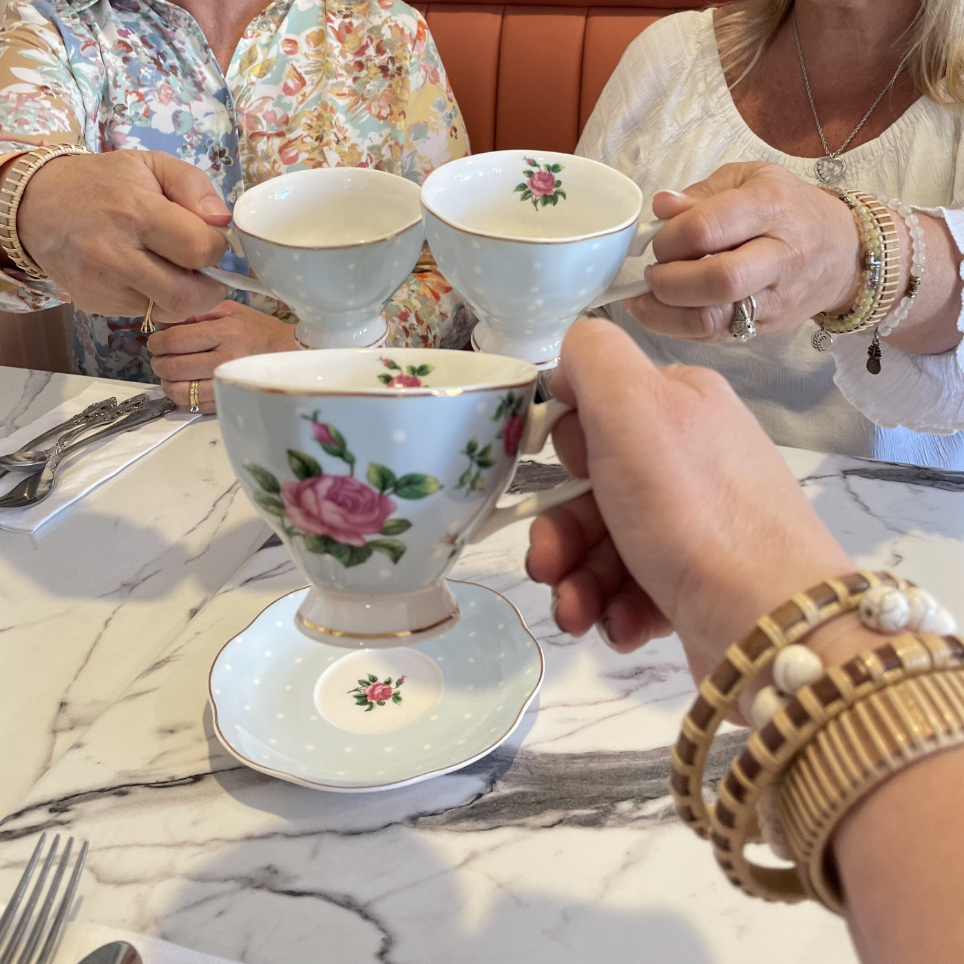 three women wearing wooden bracelets in stacks clicking floral teacups in the center of a table