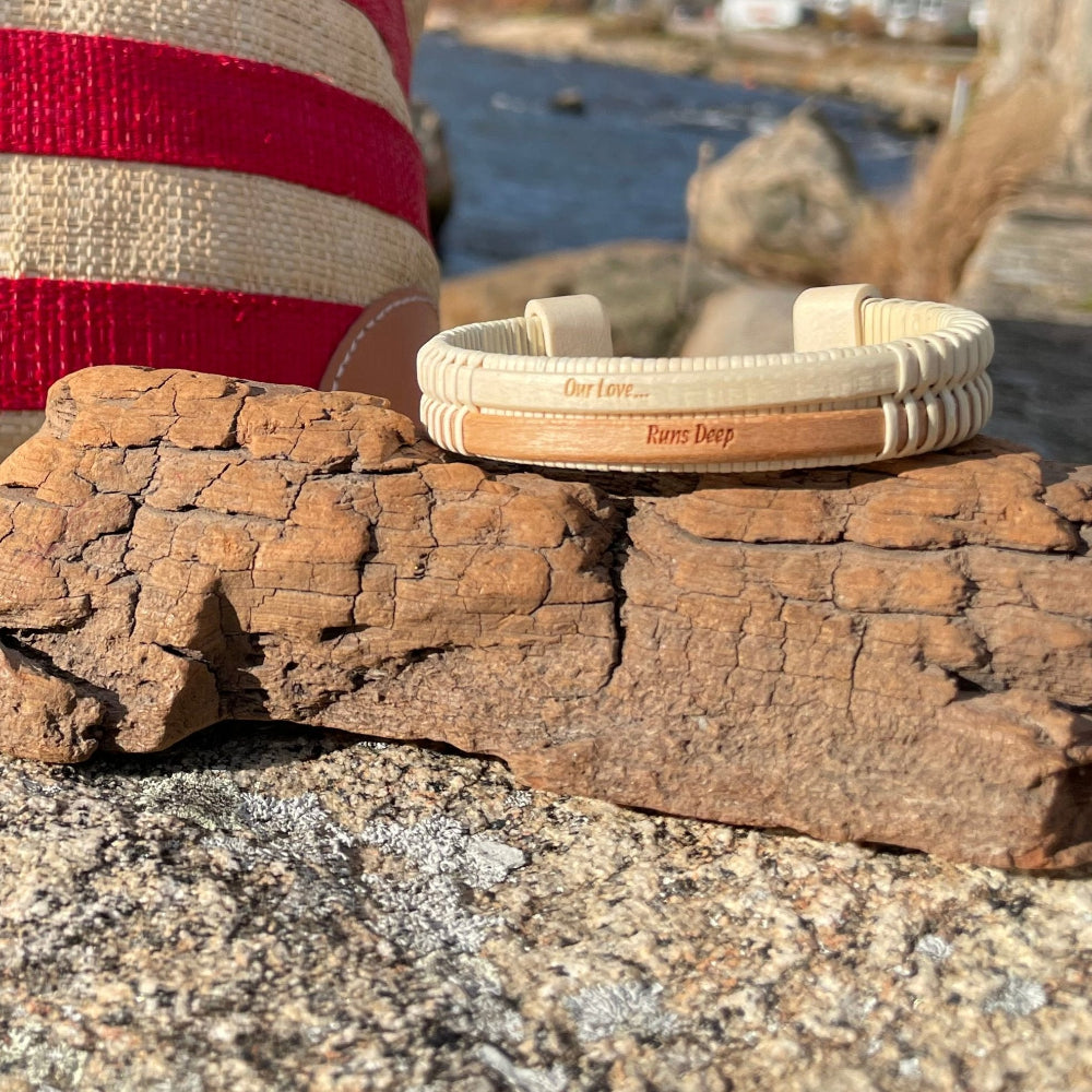 A double cuff bracelet with an etched phrase 'our love runs deep' on a wooden surface, with a red and white striped hat in the background.