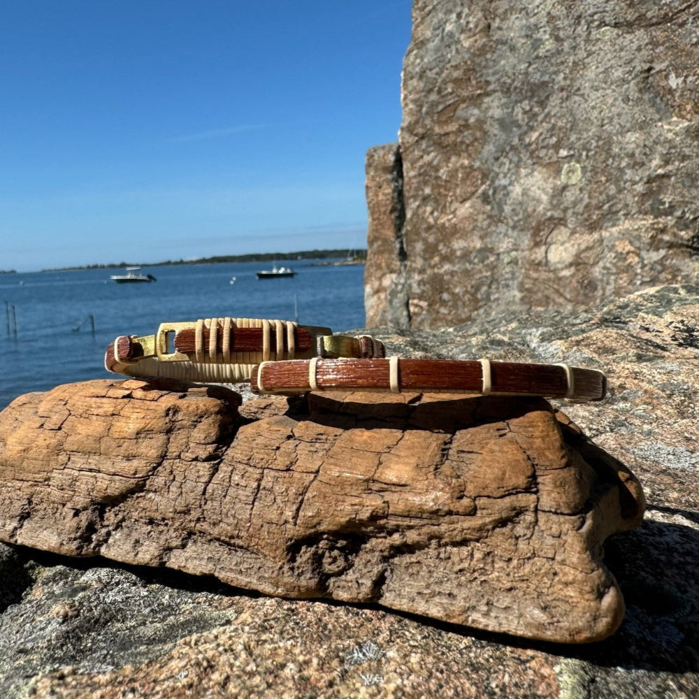 deep red wood bracelets balanced on driftwood and in front of a rock with the ocean and boats behind
