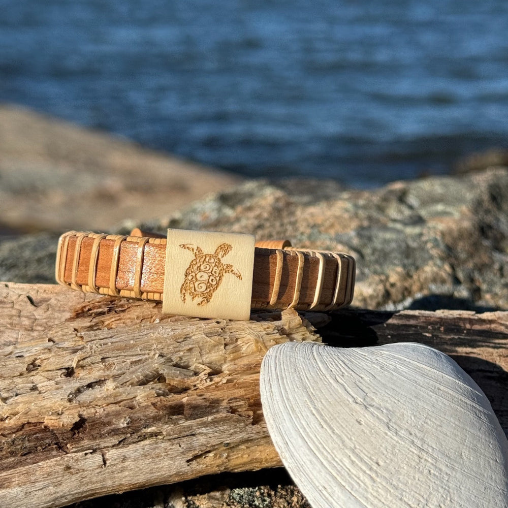 A two-tone (tan and white) wooden cuff bracelet etched with a sea turtle design, displayed on a wooden log with a beach background.
