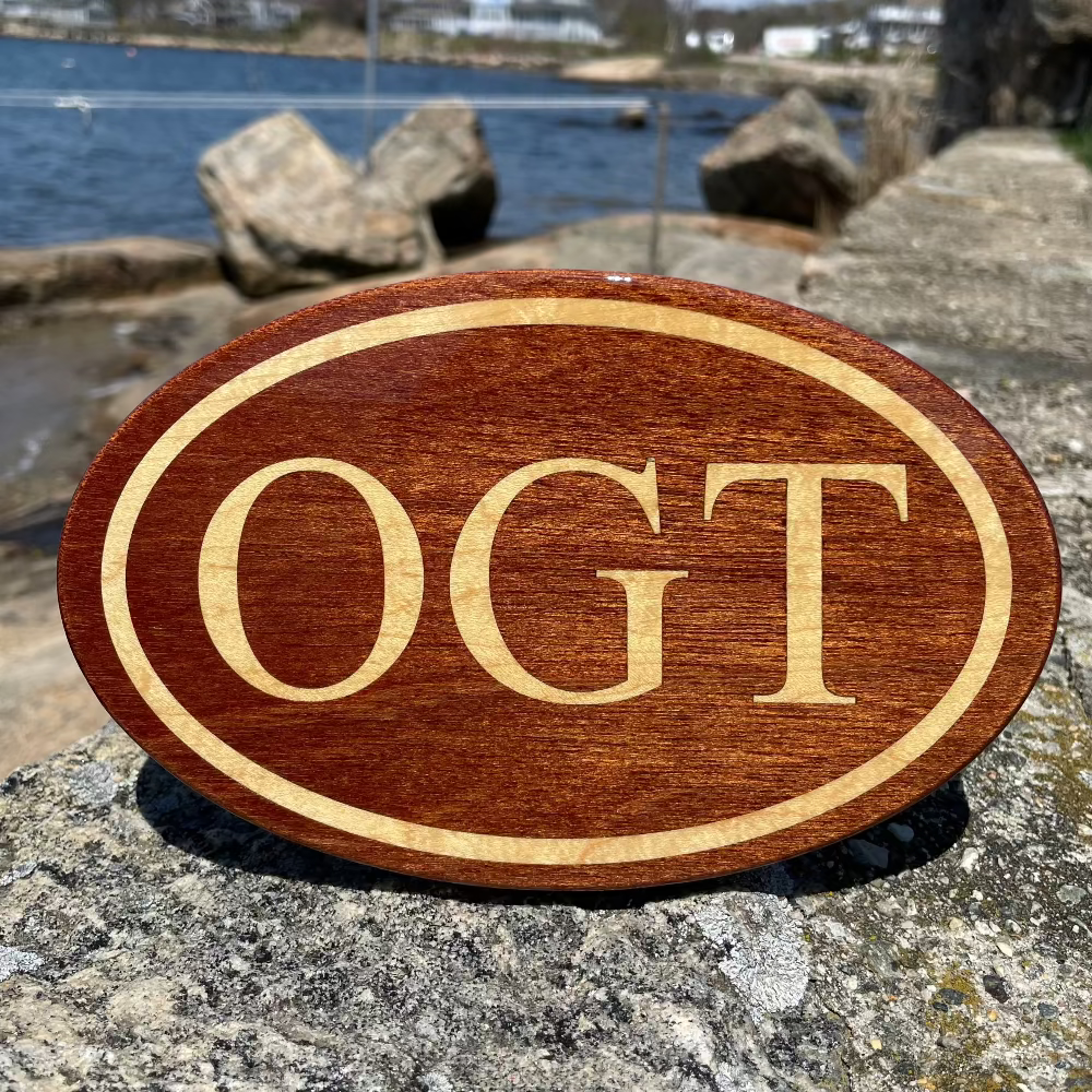A wooden hitch-cover with the letters 'OGT' carved on it, placed on a rocky surface with a body of water and sky in the background.