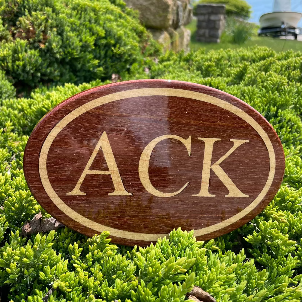 A wooden hitch cover with the initial 'A' in the center, surrounded by greenery and a stone wall in the background.