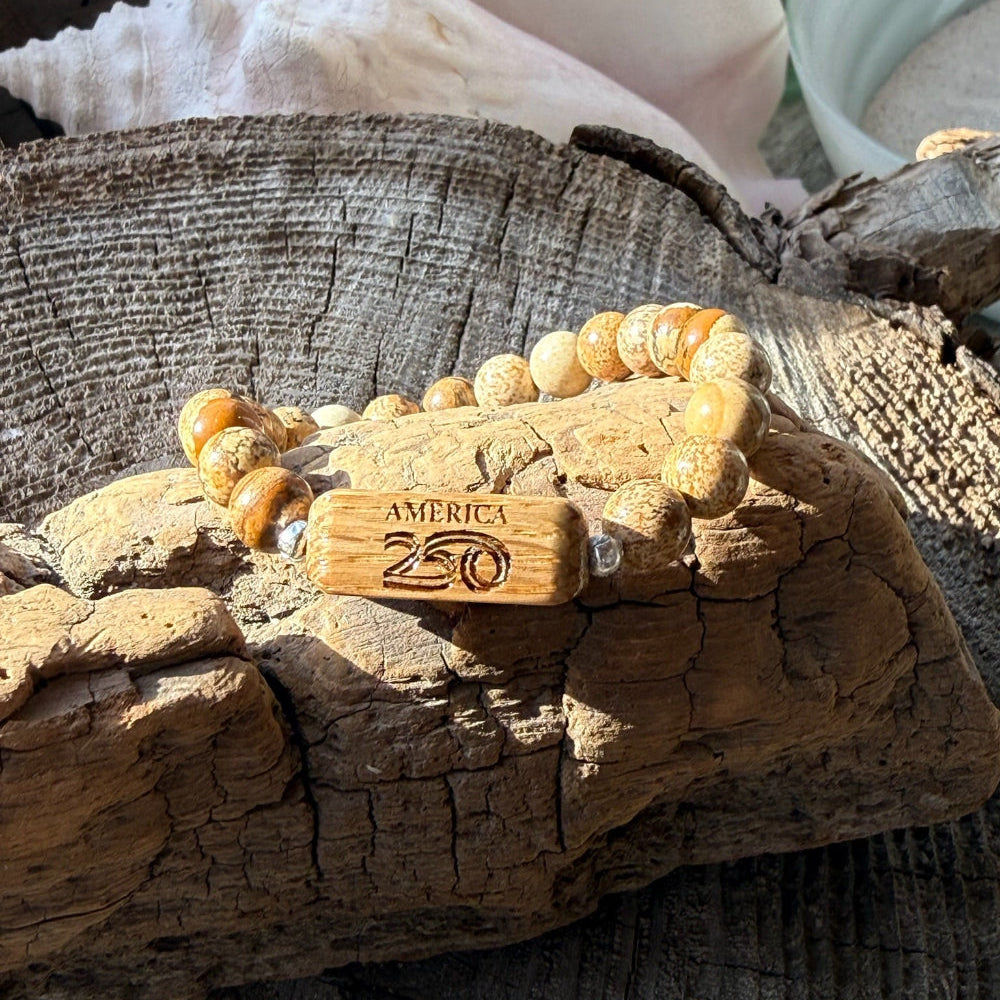 Handcrafted Old Glory Bead Bracelet resting on driftwood in sunlight, with the white oak America250 etched bead prominently displayed among Picasso jasper stone beads, made by military spouse artisans at Stonington Designs.