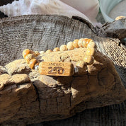 Handcrafted Old Glory Bead Bracelet resting on driftwood in sunlight, with the white oak America250 etched bead prominently displayed among Picasso jasper stone beads, made by military spouse artisans at Stonington Designs.