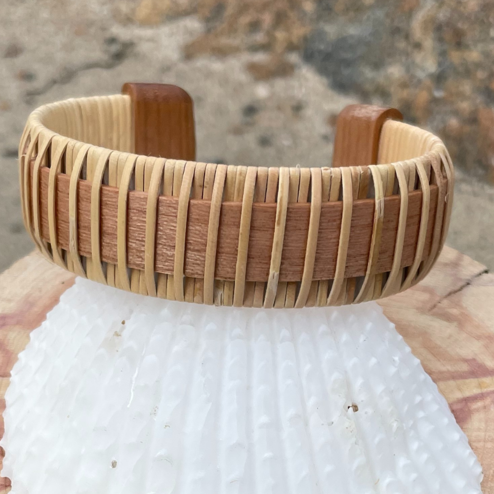 A two-tone cuff bracelet made of wood and metal, displayed on a sandy beach with a pier in the background.