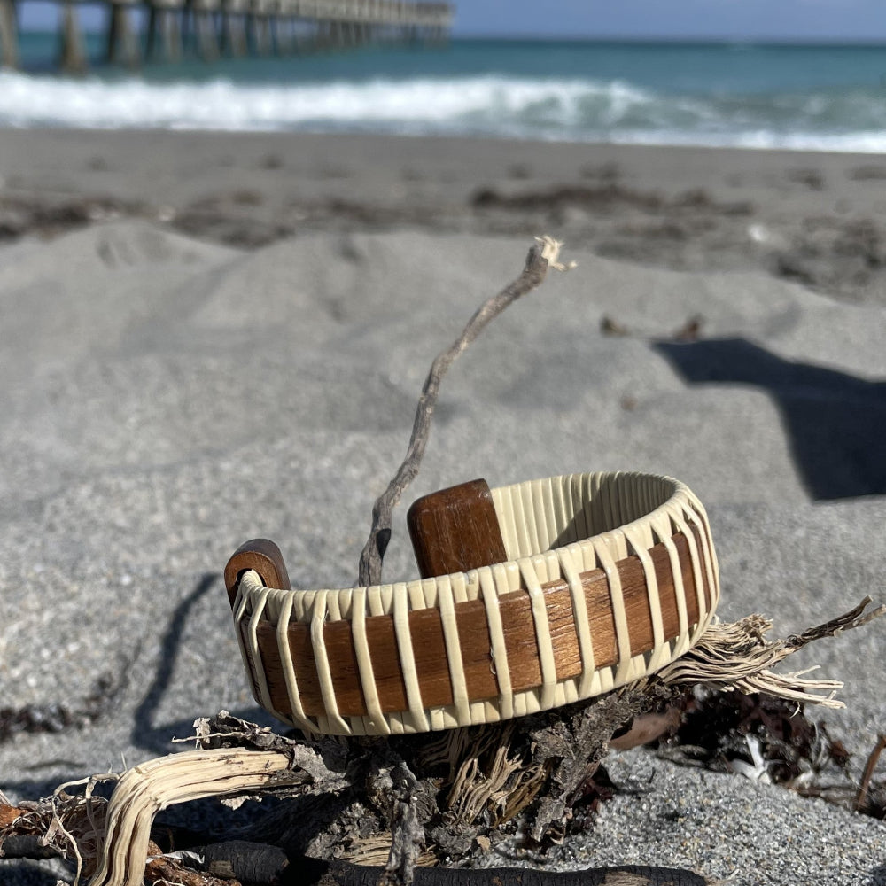 A two-tone cuff bracelet made of wood and metal, displayed on a sandy beach with a pier in the background.