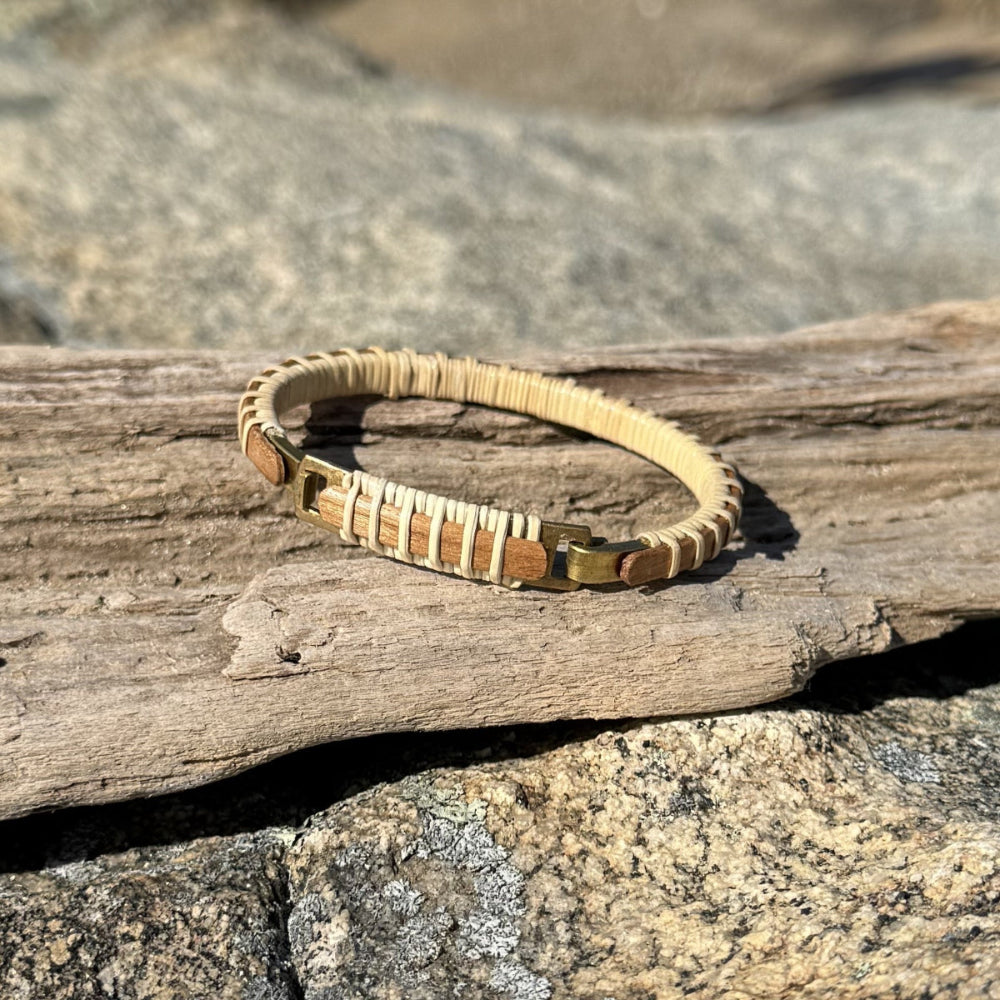 A two-tone Nauti Clasp Bracelet made of metal and wood, displayed on a wooden surface with water in the background.