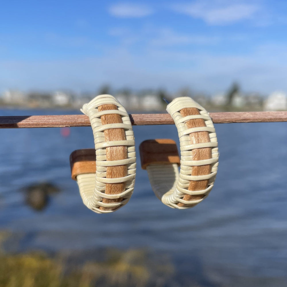 A pair of medium-sized hoop earrings made with woven design, displayed on a wooden stick, with water and buildings in the background.