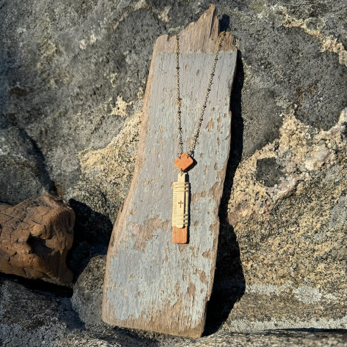 vertial woven necklace with etched cross attached to a chain and draped over a piece of driftwood