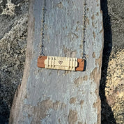 A horizontal bar necklace with a heart shape in the center, made from wood remnants, displayed on a stone surface.