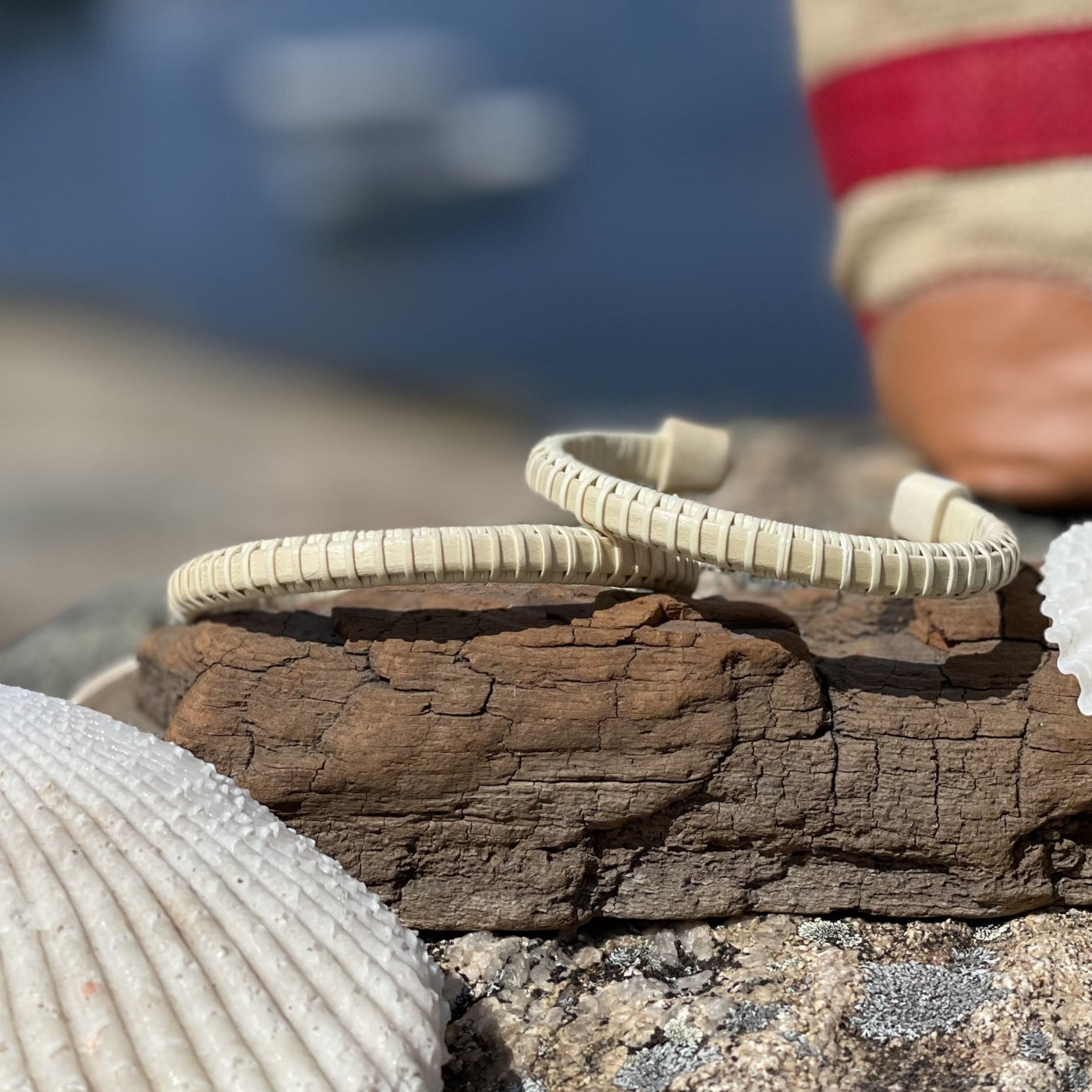 two Holly wood bracelets for women balancing on a piece of driftwood with a shell in foreground and red and rattan bag in the far corner