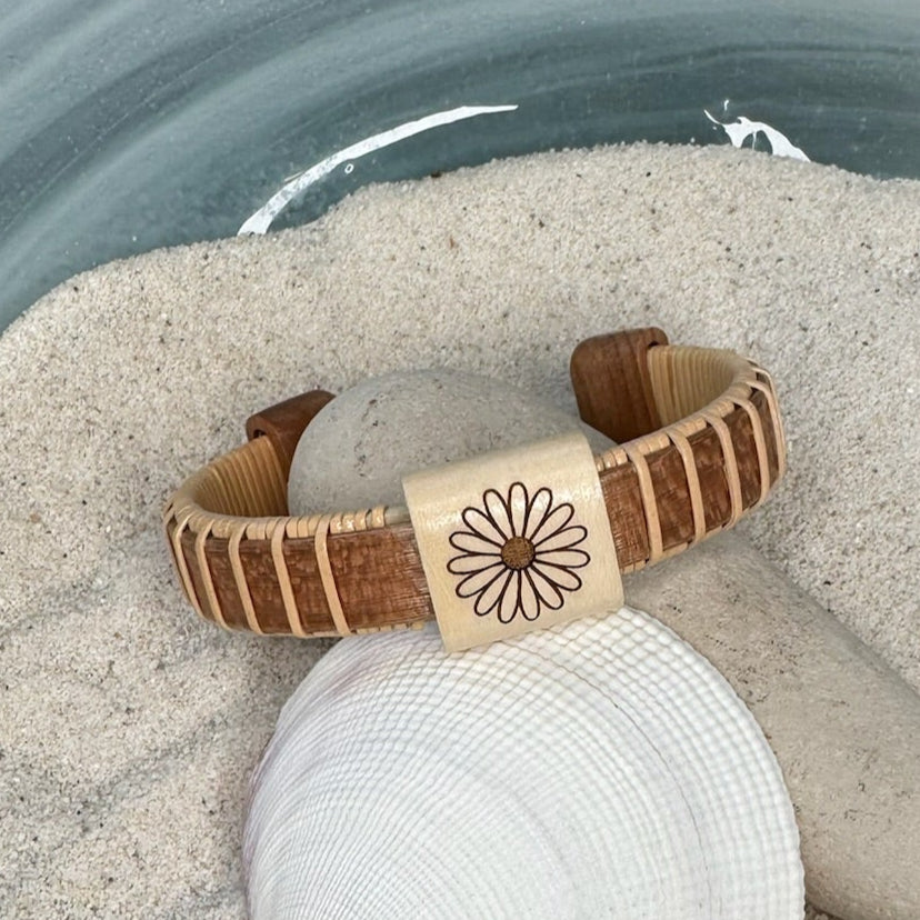 Cherry Daisy Etched cuff bracelet with a holly bead engraved with a daisy, resting on a seashell and smooth stone in sand.
