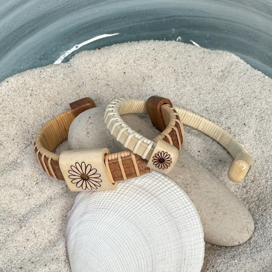 Two handcrafted Little Daisies Etched Cuff Bracelets in cherry and holly displayed together on smooth stones and a seashell in sand, showing both the slider bead and end cap daisy etching styles.