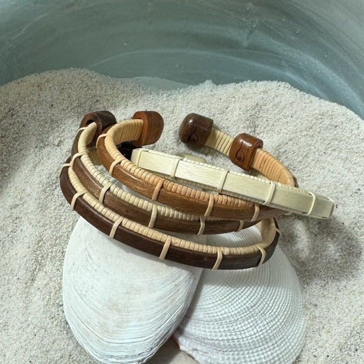 Four Bannister cuff bracelets in cherry, holly, walnut, and rosewood displayed on seashells in sand, showing all available wood variations.