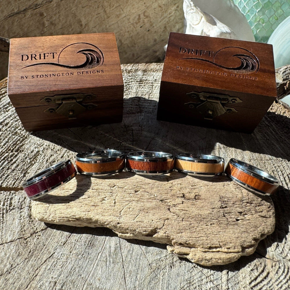 Five Beacon 8mm tungsten rings with cherry, mahogany, purple heart, rosewood, and white oak inlays displayed on driftwood alongside two engraved gift boxes.