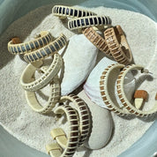 Handcrafted wood and rattan large hoop earrings in assorted wood tones scattered in sand with seashells and stones.