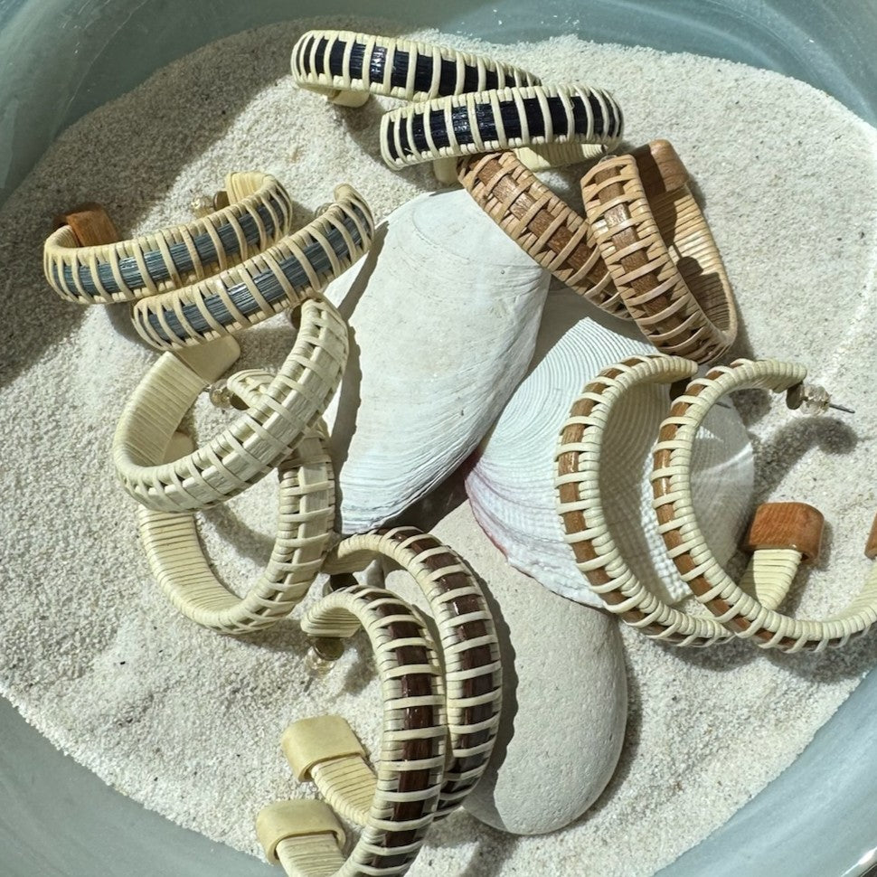 Handcrafted wood and rattan large hoop earrings in assorted wood tones scattered in sand with seashells and stones.