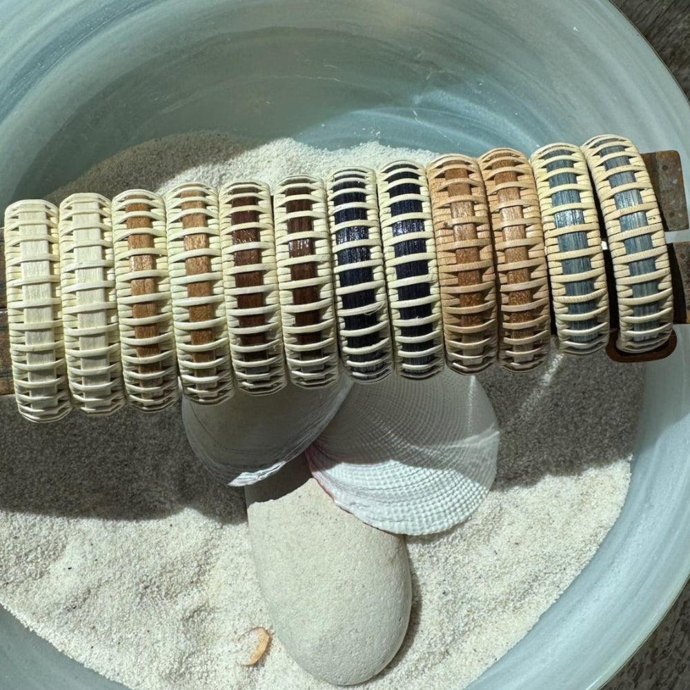 Six pairs of handcrafted large hoop earrings in a row showing the full range of wood variants, displayed in a bowl with sand, stones, and a shell.