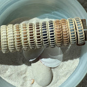 Six pairs of handcrafted large hoop earrings in a row showing the full range of wood variants, displayed in a bowl with sand, stones, and a shell.