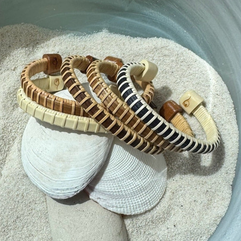 Five handcrafted Symphony cuff bracelets in cherry, holly, walnut, rosewood and navy cane with etched monogram end caps, resting in sand with seashells.