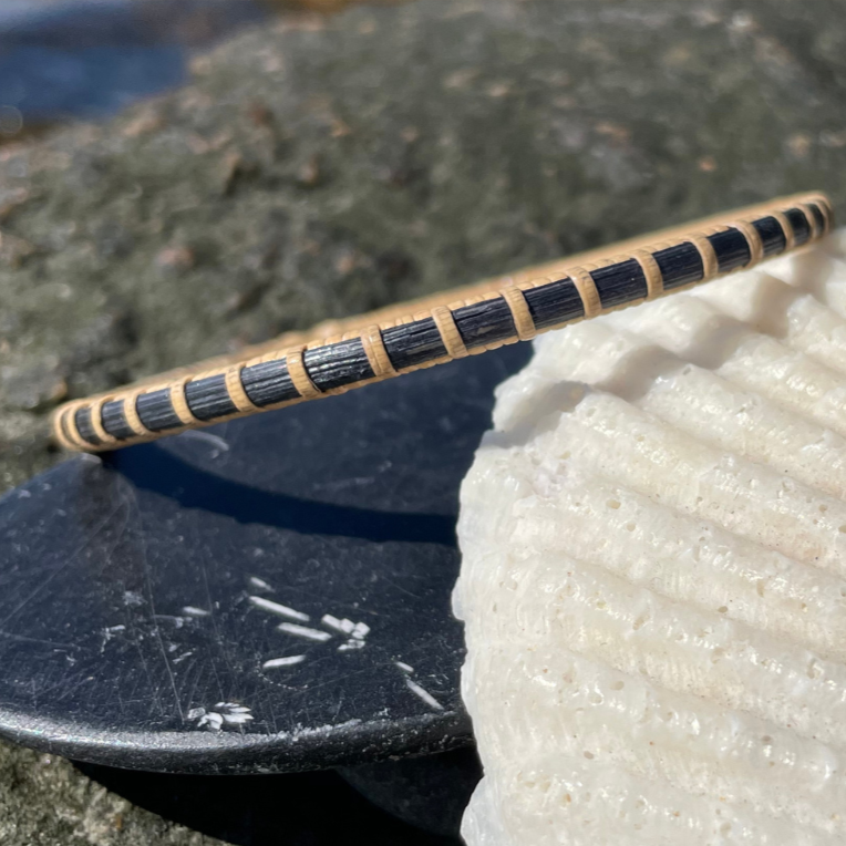 Handcrafted Ocean deep blue hand-dyed bamboo cane Secrets bangle bracelet resting on rocks beside a seashell — handmade by Military Spouse artisans in Stonington, Connecticut using American-made materials.
