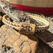Two handcrafted hand-dyed bamboo Old Glory Bangles in faded red and blue connected by a white oak slider bead etched with the America250 logo, resting on driftwood and coastal rocks, part of the licensed America250 Collection by Stonington Designs.