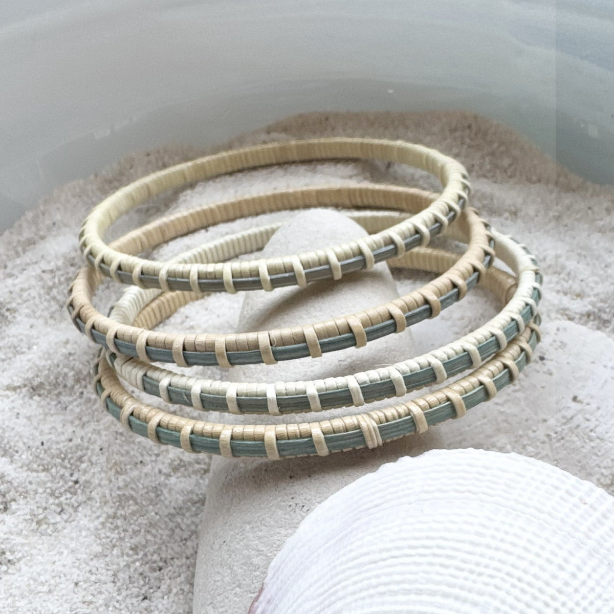 Four handcrafted hand-dyed blue bamboo cane bangle bracelets with cream woven binding, resting in sand with a shell.