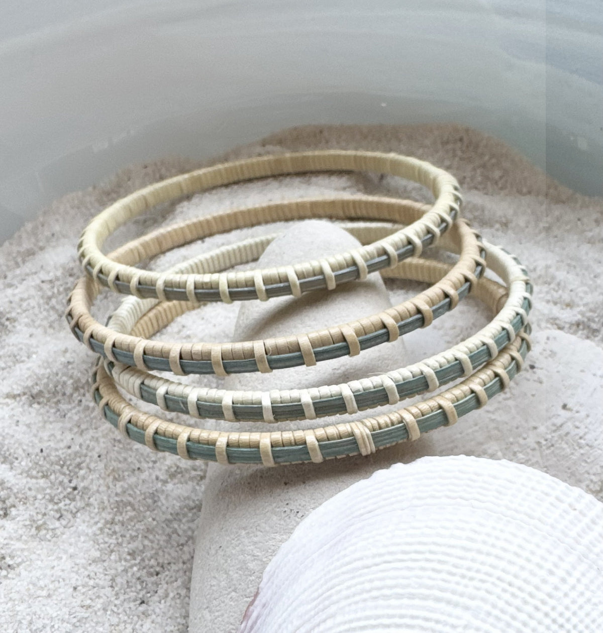 Four handcrafted hand-dyed blue bamboo cane bangle bracelets with cream woven binding, resting in sand with a shell.