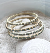 Four handcrafted hand-dyed blue bamboo cane bangle bracelets with cream woven binding, resting in sand with a shell.