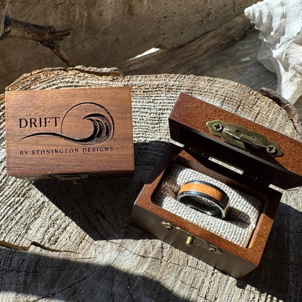 Beacon 8mm tungsten ring with cherry wood inlay shown in an open gift box beside a closed engraved Drift by Stonington Designs wooden box on driftwood.