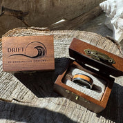 Beacon 8mm tungsten ring with cherry wood inlay shown in an open gift box beside a closed engraved Drift by Stonington Designs wooden box on driftwood.