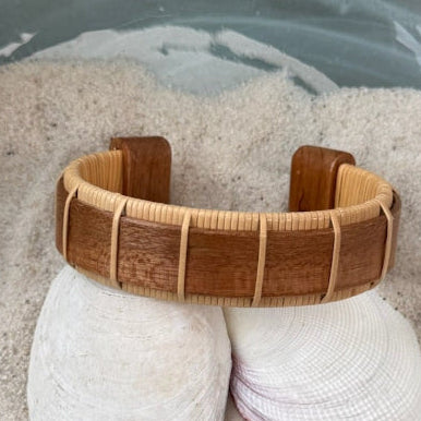 Cherry Beam cuff bracelet resting on seashells and a smooth stone in sand, showing the bold open-weave cane design.