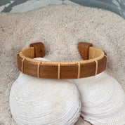 Cherry Boardwalk cuff bracelet resting on seashells and a smooth stone in sand, showing the open-weave cane design and warm wood grain.