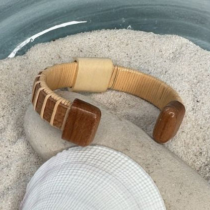 Cherry cuff bracelet with a holly bead, resting on a smooth stone and seashell in sand, showing the personalization option.