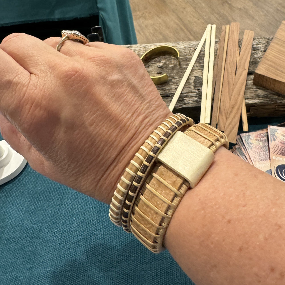 Handcrafted cherry Island Cuff Bracelet with holly slider bead worn on a wrist alongside a stacked cuff bracelet, with raw wood materials visible in the background in the studio.