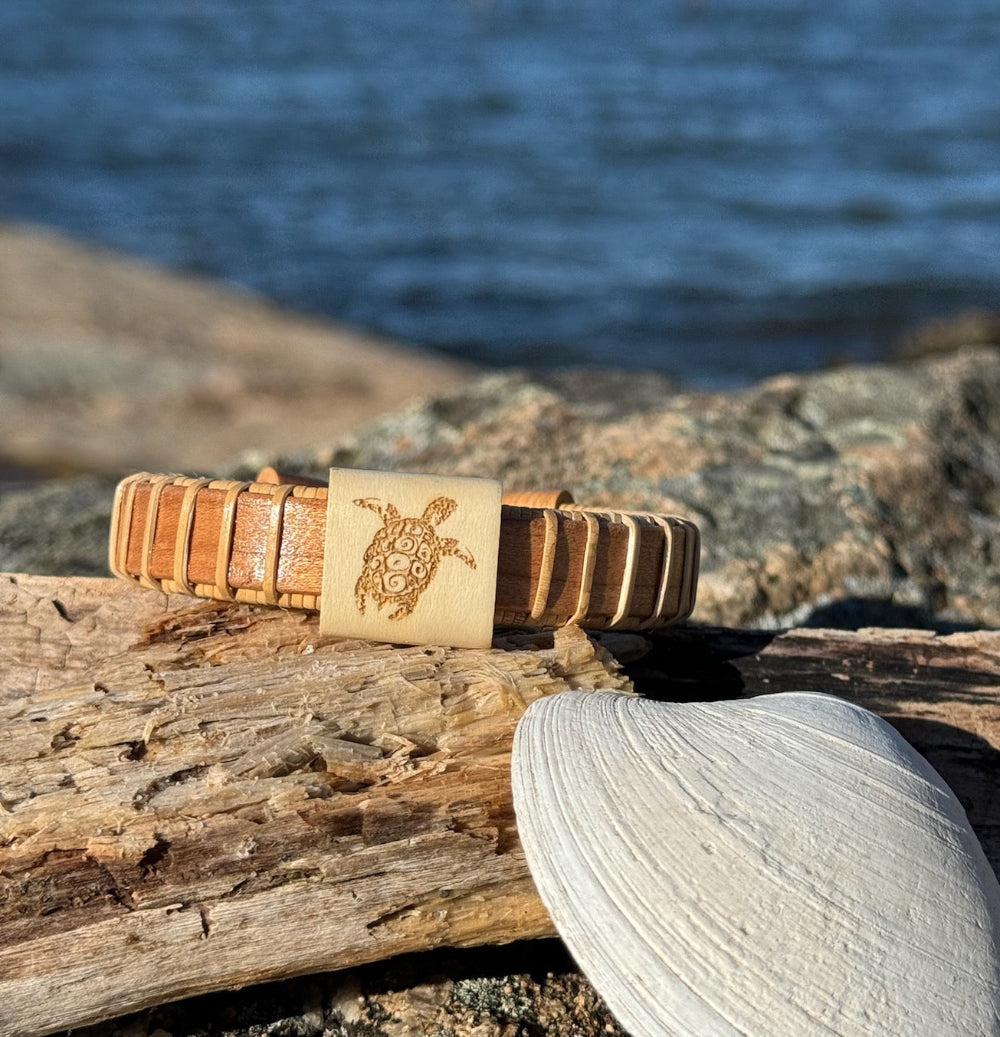 Handcrafted cherry Island Turtle Etched Cuff Bracelet with a holly slider bead engraved with a sea turtle design, resting on driftwood with a seashell and coastal water in the background, made by military spouse artisans at Stonington Designs.