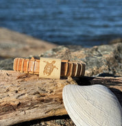 Handcrafted cherry Island Turtle Etched Cuff Bracelet with a holly slider bead engraved with a sea turtle design, resting on driftwood with a seashell and coastal water in the background, made by military spouse artisans at Stonington Designs.