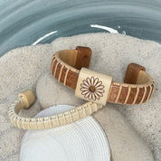 Handcrafted cherry Little Daisies Etched Cuff Bracelet with a large holly slider bead engraved with a daisy motif, draped over smooth stones and a seashell in sand.