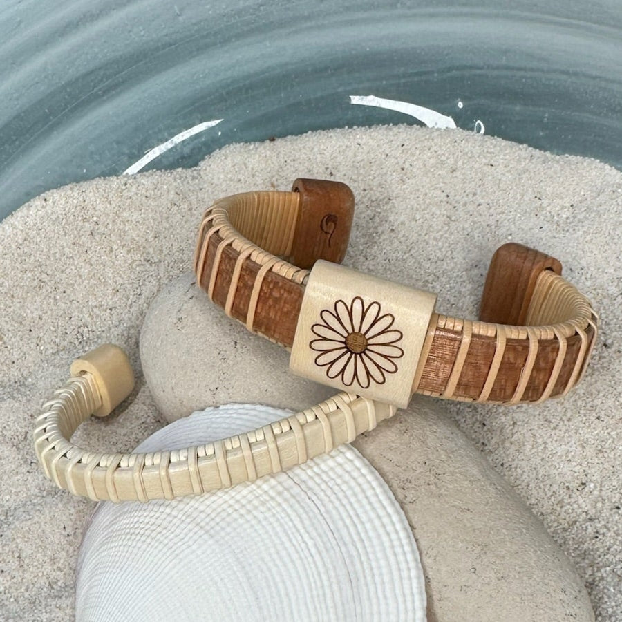 Handcrafted cherry Little Daisies Etched Cuff Bracelet with a large holly slider bead engraved with a daisy motif, draped over smooth stones and a seashell in sand.