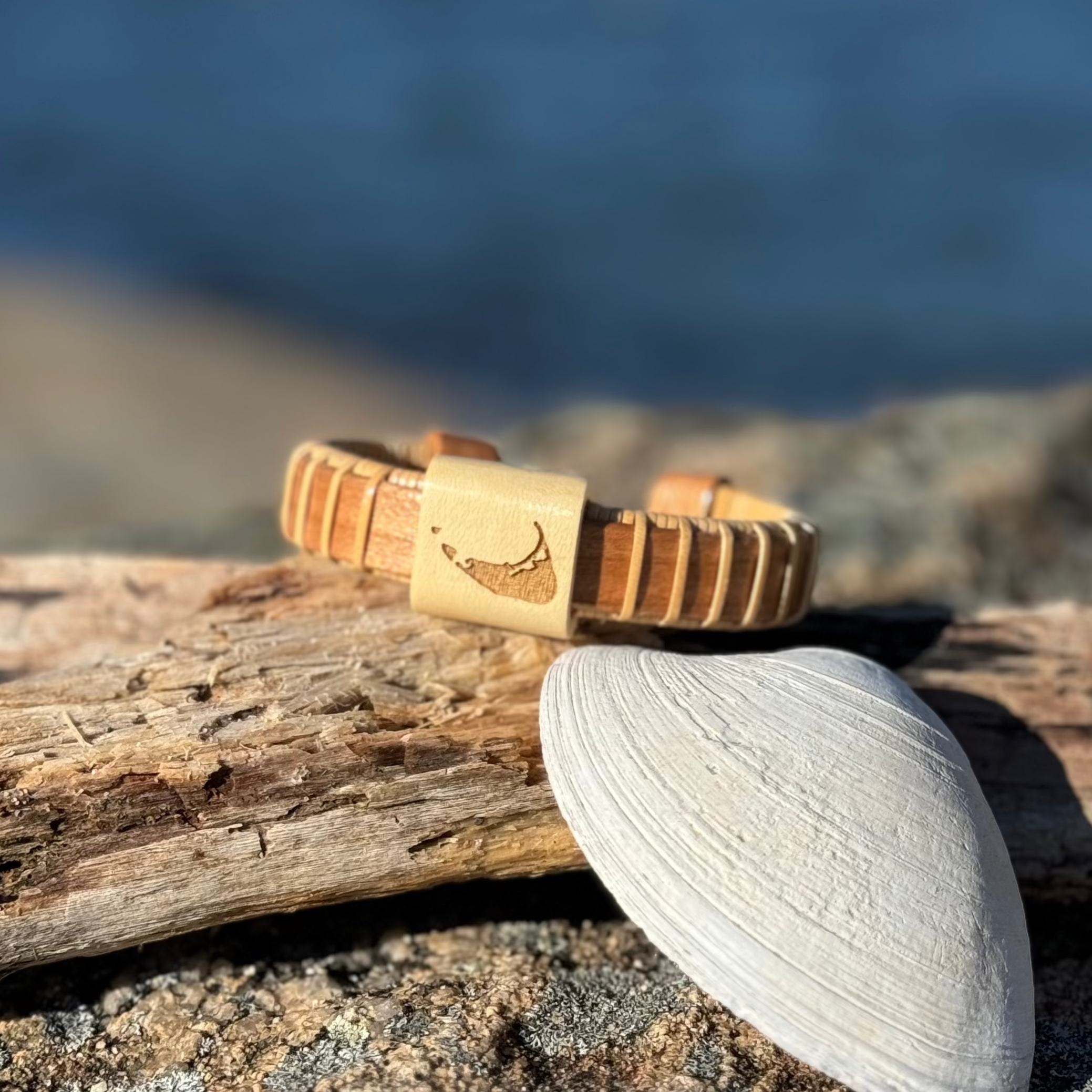 Handcrafted cherry wood cuff bracelet with an etched Nantucket Island shape on the holly wood center bead, resting on driftwood near the water with a shell.