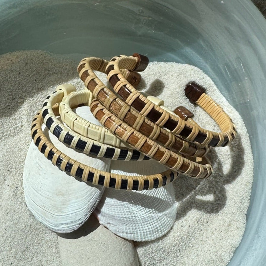 Five Clubhouse cuff bracelets in cherry, holly, navy cane, rosewood, and walnut displayed on seashells in sand, showing the open-weave cane design and available wood variations.
