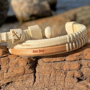 Close-up of the cherry and holly Our Love Runs Deep Etched Double Cuff Bracelet resting on driftwood, showing the anchor-engraved end cap, holly bead detail, and "Our Love... Runs Deep" text etched on the cherry stave.