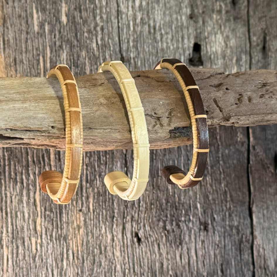 Three Bannister cuff bracelets in cherry, holly, and rosewood displayed on driftwood, showing the range of available wood tones.