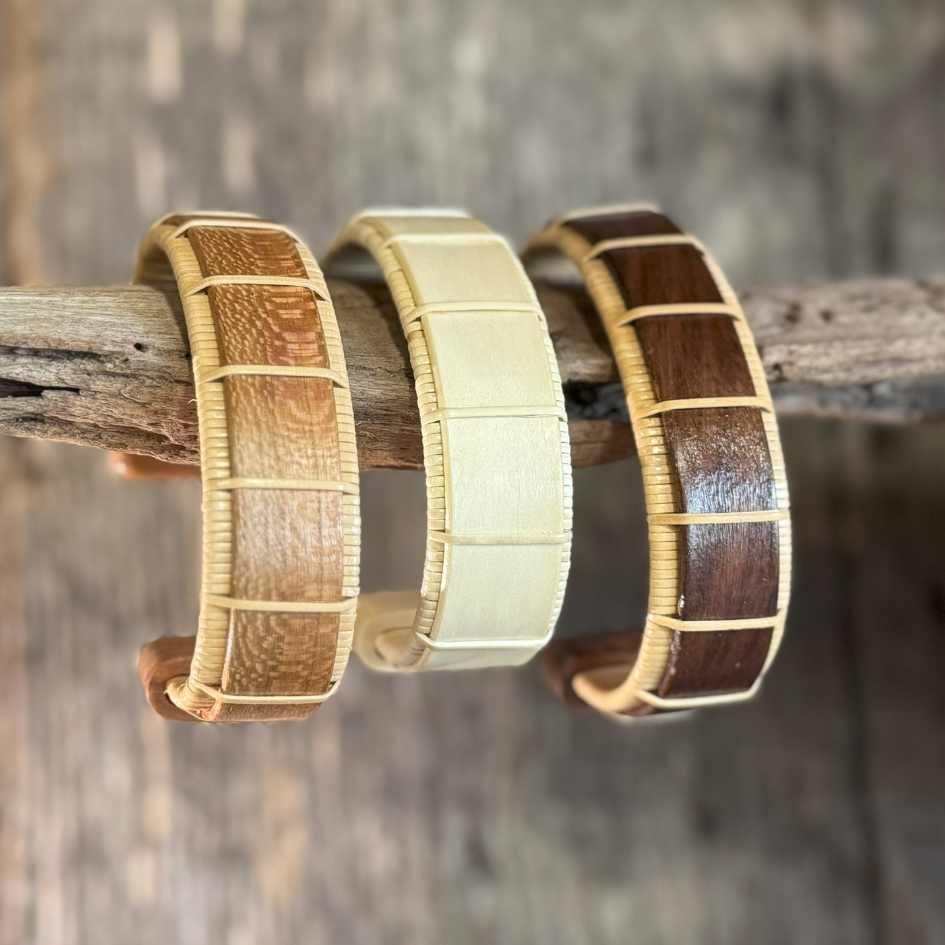 Three Beam cuff bracelets in cherry, holly, and rosewood hanging on a piece of driftwood, showing the wide open-weave cane design and wood grain detail.