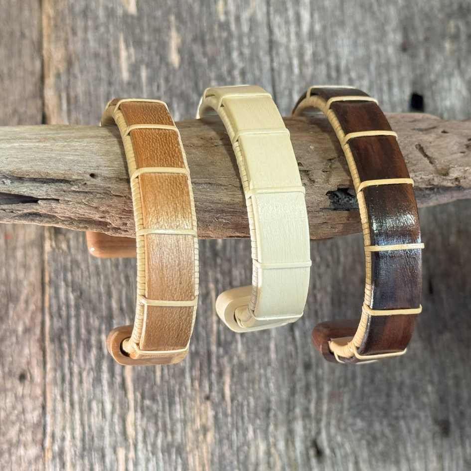 Three Boardwalk cuff bracelets in cherry, holly, and rosewood hanging on a piece of driftwood, showing the wide open-weave cane design and wood grain detail.