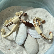 Multiple Cove cuff bracelets in cherry, holly, and rosewood with matching sliders displayed on seashells in sand.