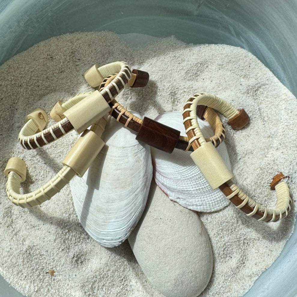 Multiple Cove cuff bracelets in cherry, holly, and rosewood with matching sliders displayed on seashells in sand.