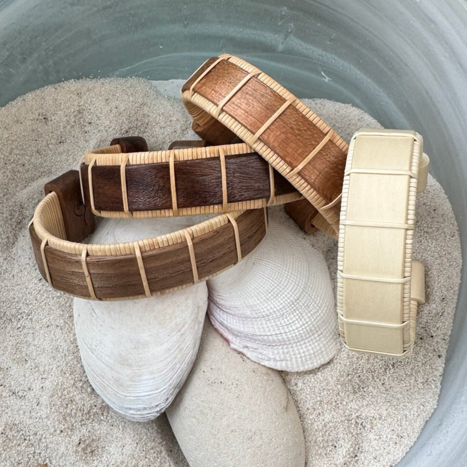 Four Beam cuff bracelets in walnut, rosewood, holly, and cherry displayed on seashells in sand, showing the bold open-weave design and available wood variations.